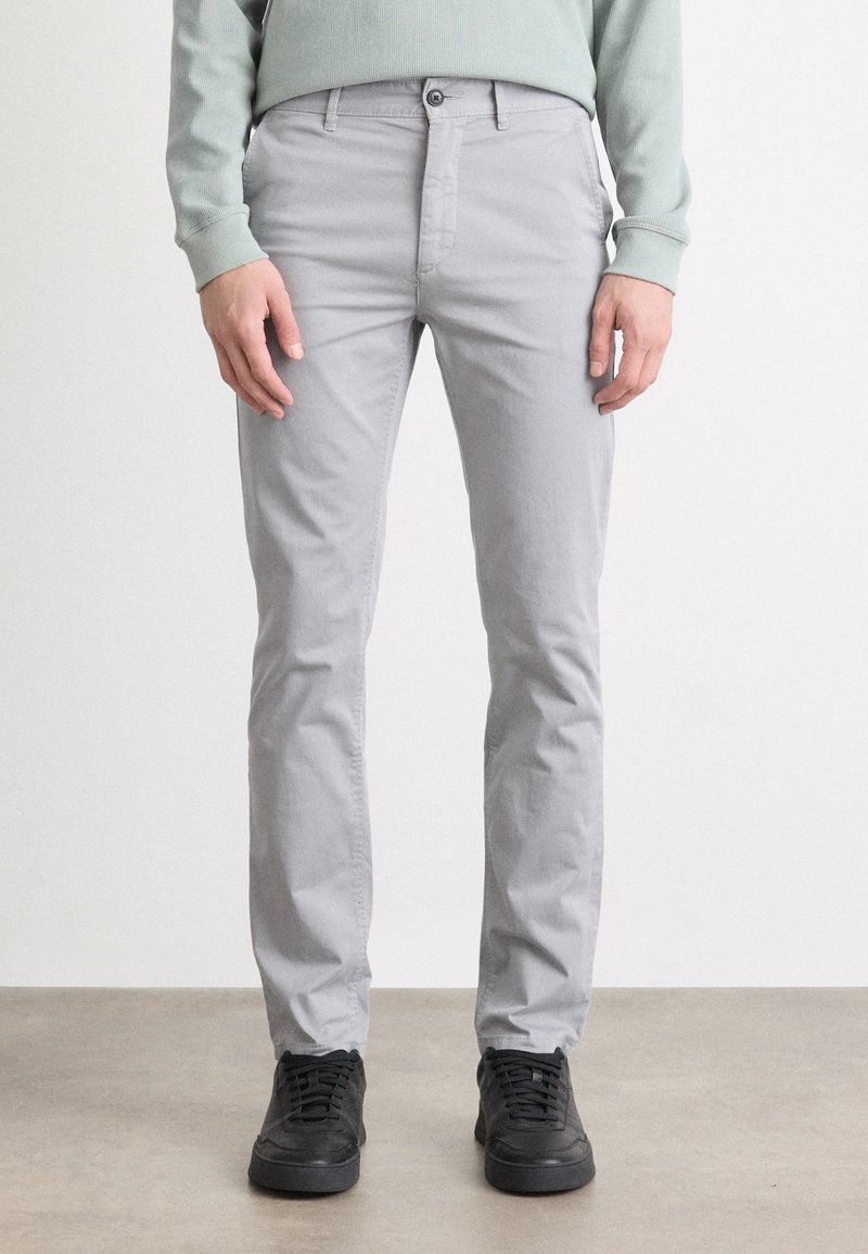 Jasnoszare chinosy o dopasowanym kroju, z zapięciem na guziki, szlufkami na pasek i kieszeniami. W zestawieniu z czarnymi trampkami na neutralnym tle.