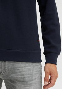 Sweatshirt texturé bleu marine avec un col et des poignets côtelés, comportant une petite étiquette de marque en bas, associé à un jean gris clair.