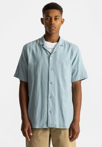 Lichtblauw overhemd met korte mouwen en een ontspannen pasvorm, met een gestructureerde stof en een kraag. Draagt over een wit T-shirt met ronde hals.