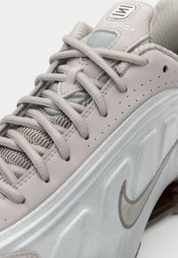Basket de sport gris clair avec un dessus en cuir lisse, des accents argent métallisé, une languette texturée et des lacets gris. Présente le logo Nike Shox.