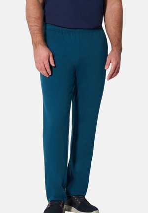 Man wearing dark teal casual pants, navy blue shirt, and black shoes standing against a plain light background.