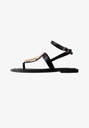 Czarna płaska sandałka z paskiem wokół kostki i złotym łańcuszkiem przy pasku między palcami, posiadająca boczne zapięcie na klamrę.