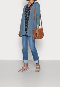 Světle modrý žebrovaný kardigan přes tmavě modrou květovanou blůzu, doplněný o ohrnuté modré džíny a světle modré otevřené sandály. Hnědá kabelka přes rameno.