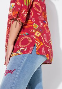 Blusa roja con patrón de corta mangas que presenta varios diseños multicolores. Llevada con jeans de color azul claro, mostrando texto bordado.