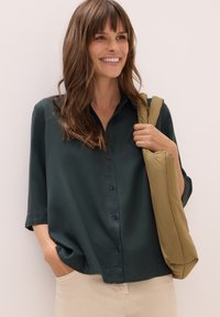 Leende kvinna med långt brunt hår som bär en mörkgrön skjorta med knappar, beige byxor och en vadderad axelväska i olivgrön färg.