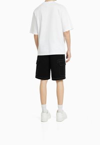 Valkoinen puuvillainen t-paita lyhyillä hihoilla, yhdistetty mustiin cargo-shortseihin, joissa on sivutaskut ja valkoinen ompelu, käytössä valkoisten lenkkarien kanssa.