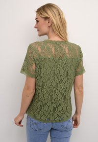 Top in pizzo verde oliva con orlo ondulato e maniche corte, caratterizzato da intricati motivi floreali e un design semi-trasparente.