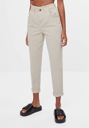 Beige Hose mit hoher Taille und Paperbag-Bund, mit umgeschlagenen Bündchen, vorderen Taschen, getragen mit schwarzen gepolsterten Slide-Sandalen an einer stehenden Person.