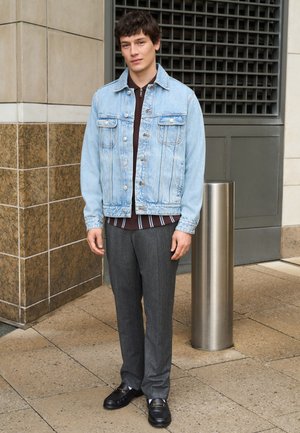 Nuori mies seisoo ulkona rakennuksen lähellä, hänellä on päällään vaaleansininen farkkutakki, raidallinen paita, harmaat housut ja mustat loafersit.