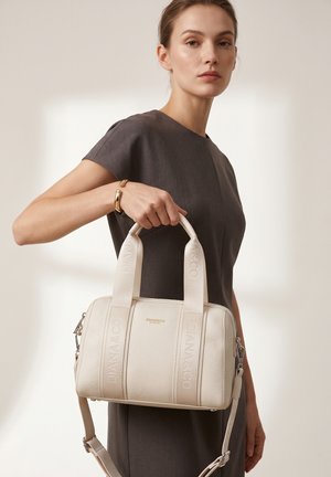 Donna in abito scuro a maniche corte che tiene una borsa beige Diana&Co con doppi manici e tracolla, indossa un braccialetto d'oro.