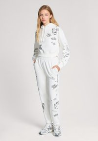 Felpa bianca con cappuccio e pantaloni jogger, caratterizzati da stampe grafiche nere, inclusi personaggi e testo, con una vestibilità rilassata e regolazioni con cordini.