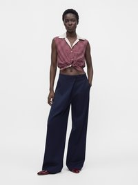Γυναίκα που φοράει αμάνικο crop top με κόκκινες και λευκές λωρίδες, σκούρο μπλε παντελόνι με φαρδιά πόδια και κόκκινα παπούτσια, στέκεται μπροστά σε απλό φωτεινό φόντο.