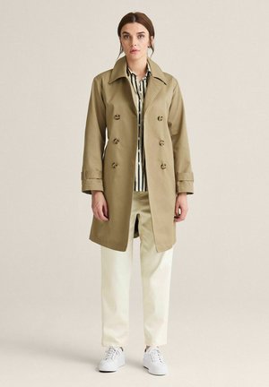Tan trenchcoat med dobbeltbrystede knapper, stor krave og løs pasform, parret med en stribet skjorte og cremfargede bukser.