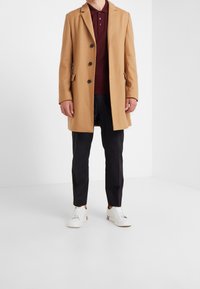 Kamelfrakke med slagkrave, enkeltknap design, fem knapper, sidelommer, båret over en bordeaux polo og sorte bukser, med hvide sneakers.