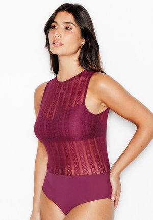 Femme aux longs cheveux foncés portant un body en dentelle bordeaux sans manches, debout avec une main sur la hanche, regardant vers la gauche sur fond blanc.