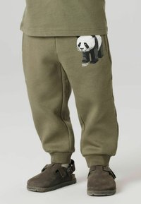 Joggebukser i olivengrønn i et mykt materiale, med et panda-motiv på lommen. Paret med brune tresko med justerbare stropper.