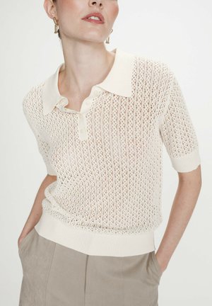 Femme portant un polo en maille crème à manches courtes avec un motif ajouré délicat et un pantalon beige taille haute, les mains dans les poches.