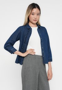 Tume denim särk volangiga ees, küljetaskud ja nööbitav kinni. Kombineeritud heledate hallide mustriliste pükste ja valge ülaosaga.