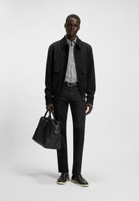 Veste bomber noire, chemise rayée, pantalon noir ajusté, chaussures en cuir noires avec semelles blanches, tenant un sac noir texturé.