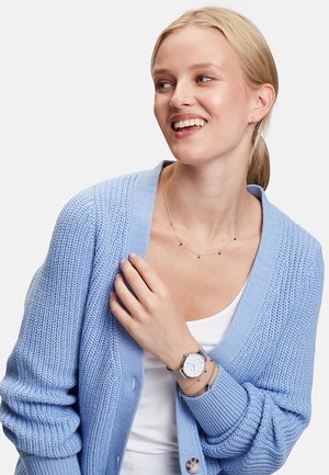 Cardigan tricoté bleu clair avec un col en V, des manches longues. Le modèle porte une montre en argent, des colliers superposés et un haut blanc. Texture lisse.