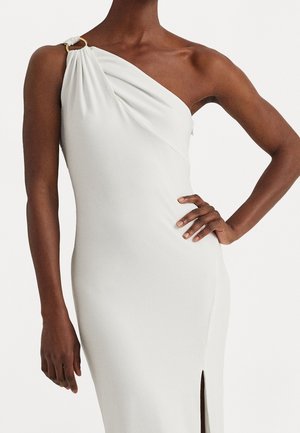 Femme portant une robe blanche sans manches à une épaule avec un détail en anneau doré et une fente haute sur la cuisse, main sur la hanche.