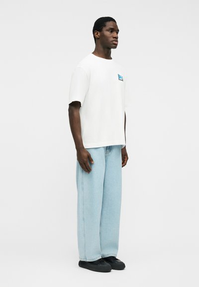 Άνδρας που στέκεται φορώντας λευκό κοντομάνικο μπλουζάκι με μικρό μπλε γραφικό και ανοιχτό μπλε παντελόνι με φαρδιά μπότα και μαύρα παπούτσια.