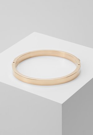 Gładka, okrągła złota bransoletka typu bangle spoczywa na białym geometrycznym piedestale z minimalistycznym szarym tłem.