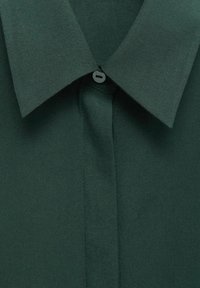 Col de chemise vert foncé avec un petit bouton noir unique fixé au centre supérieur de la patte de boutonnage.