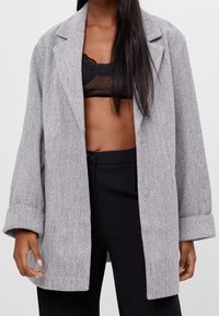 Grauer Fischgrätmantel mit Reverskragen und aufgekrempelten Ärmeln, aus strukturiertem Stoff, getragen über einem schwarzen Netz-Oberteil und schwarzen Hosen.