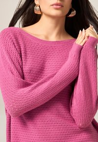 Luźny sweter w jasnoróżowym kolorze, z fakturowanym wzorem dzianiny, szerokim dekoltem i długimi rękawami z prążkowanymi mankietami.