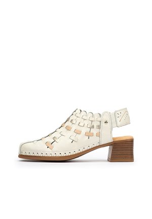 Zueco de cuero blanco con tacón, diseño trenzado, parte trasera abierta y tacón de madera marrón claro. Presenta acentos beige y metálicos en la trenza.