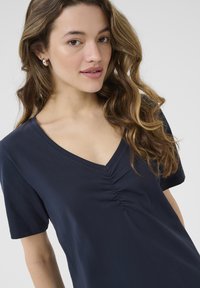 Haut bleu marine à manches courtes avec un décolleté en V et un détail de ruché à l'avant. Fabriqué en tissu doux, offrant un design ajusté et décontracté.