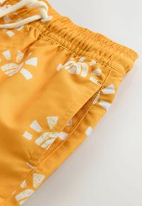 Sárga fürdőnadrág fehér geometriai mintával, rugalmas derékpánttal és húzózsinórral, valamint oldalzseb részlettel. Sima szövet textúra.