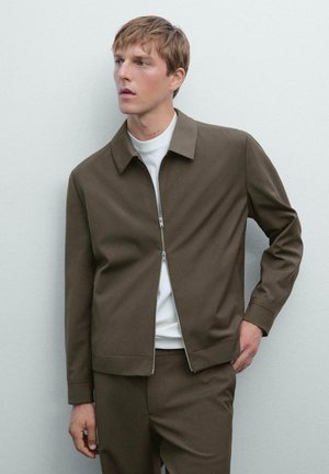 Brown zip-up jacket made of textured fabric, featuring a collared neck and a simple design. Worn over a white shirt.