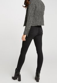 Svarta tajta jeans har hög midja med knappdetaljer längs det yttre benet och är kombinerade med en strukturerad svart och vit jacka.