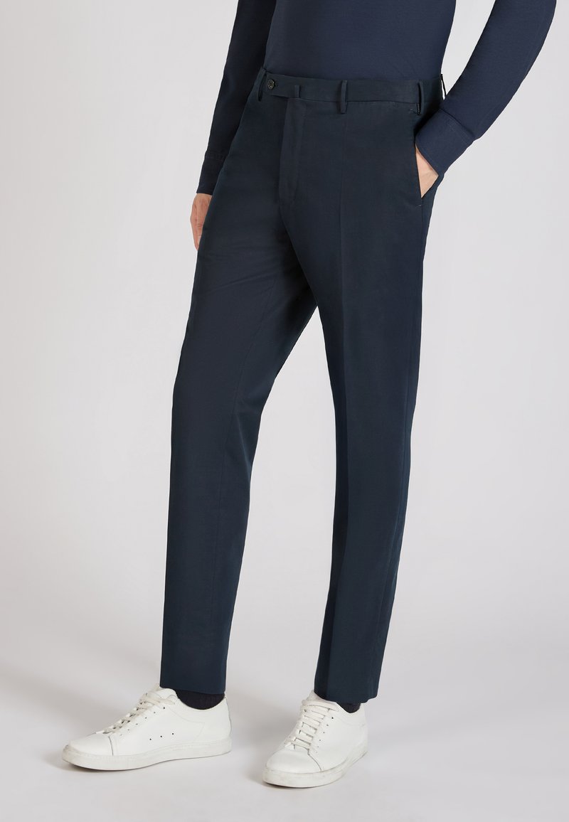 Pantalon marine en tissu lisse, avec une coupe ajustée, des poches avant et une fermeture à un bouton. Associé à des baskets blanches.