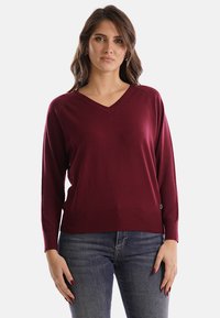 Donna che indossa un maglione bordeaux a maniche lunghe con scollo a V e jeans blu, in piedi con le mani rilassate lungo i fianchi davanti a uno sfondo neutro.