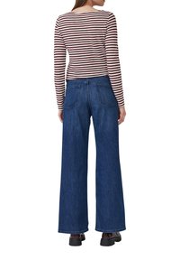 Vrouw in hoge taille wijde blauwe jeans met achterzakken, een lange mouwen top met bruine en witte strepen, en donkere schoenen, staande met haar gezicht naar de achterkant.