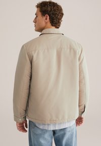 Beige, gewatteerde jas met een kraag, drukknoopaccenten en zichtbare stiksels. Draagover een gestreept shirt en blauwe spijkerbroek. Ontspannen pasvorm.