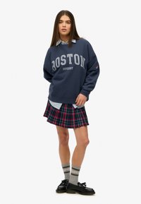 Sudadera azul marino con estampado de "BOSTON", superpuesta sobre una camisa de collar azul claro, combinada con una falda de cuadros, calcetas grises y zapatos negros.