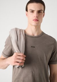 Szürke pamut póló kerek nyakkal és tónusos logóval, egy olyan személyen, aki egy világos bézs pulóvert tart a válla felett.