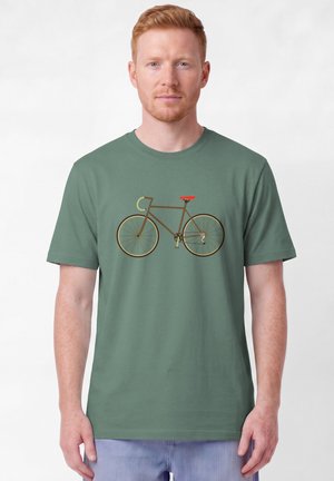 Mann mit kurzen roten Haaren, der ein grünes T-Shirt mit einer braunen Fahrradgrafik mit rotem Sattel und hellbraunen Lenker trägt.