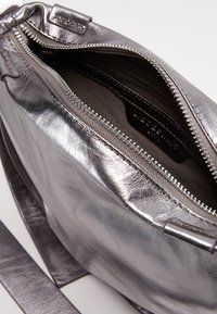 Metallinen hopeinen käsilaukku, jossa on sileä pinta, vetoketjukiinnitys ja kankainen vuori. Varustettu säädettävällä hihnalla ja minimalistisella muotoilulla.