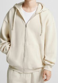 Felpa crema con zip, dotata di ampie tasche frontali, cappuccio regolabile con cordino e polsini a coste; realizzata in morbido tessuto testurizzato.
