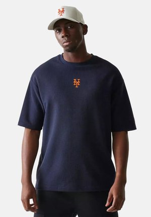Hombre con camisa de manga corta azul marino y gorra gris claro, ambas con el logo "NY" naranja en el frente, de pie contra un fondo blanco liso.