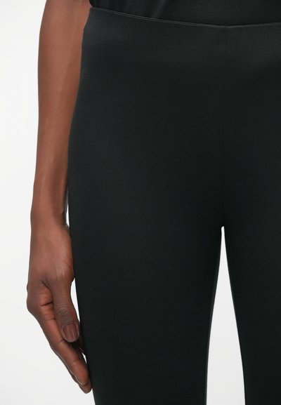 Zwarte leggings met een gladde textuur en een hoogwaardig ontwerp. Close-up view toont een hand die tegen de zijnaad rust.