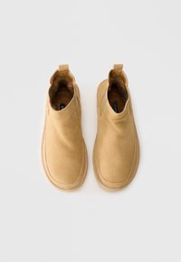 Une paire de bottines à enfiler en daim beige avec doublure en fourrure, pointes arrondies et languettes, sur fond blanc.
