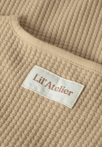 Tejido de punto de gofre beige con escote redondeado, que presenta una etiqueta blanca con la inscripción "Lil'Atelier" en letras marrones.