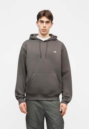 Νέος άνδρας που φοράει απλό σκουρόγκρι φούτερ με κουκούλα, εμπρόσθια τσέπη και λογότυπο στο στήθος, συνδυασμένο με σκουρόγκρι παντελόνι καμπαρντίνας, στέκεται μπροστά σε λευκό φόντο.