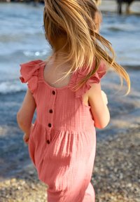 Rosa Baumwoll-Jumpsuit mit Rüschennähten und Knopfdetails. Der Stoff hat eine strukturierte Oberfläche und das Design verfügt über einen elastischen Bund.